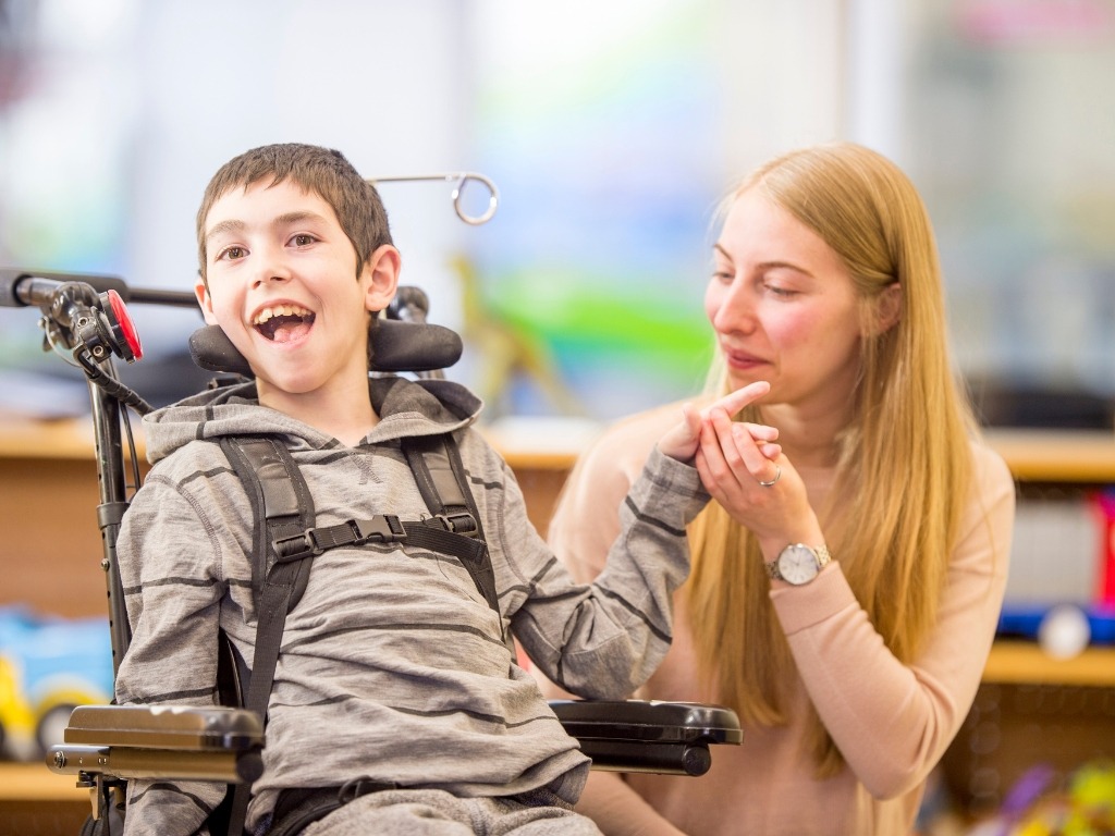 Child in a wheelchair smiling with teacher in a classroom.