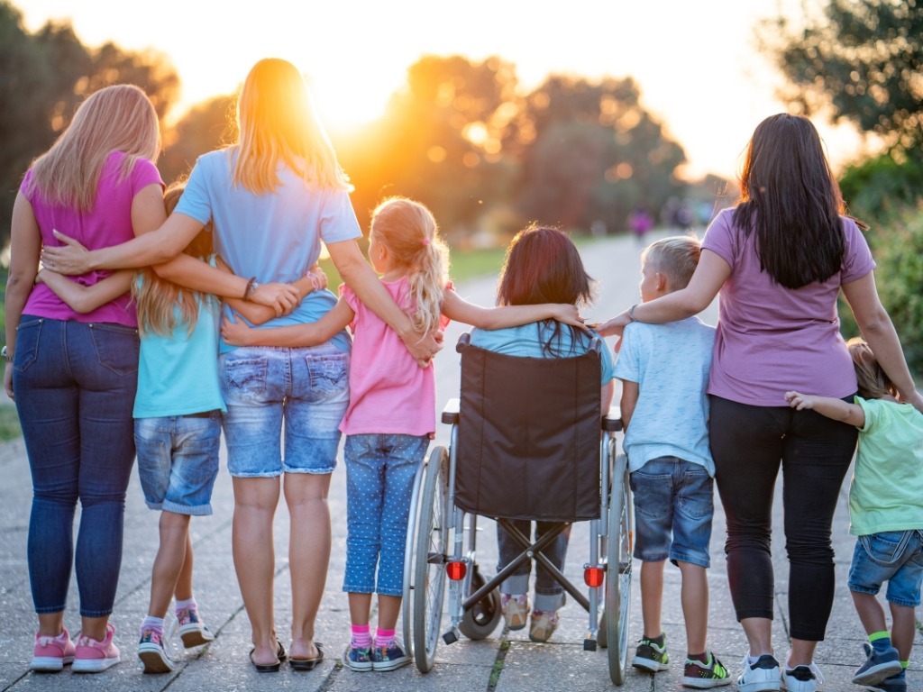 3 adults and 5 children (one child is sitting in a wheelchair) are all hugging with their backs to the camera and watching the sunset.