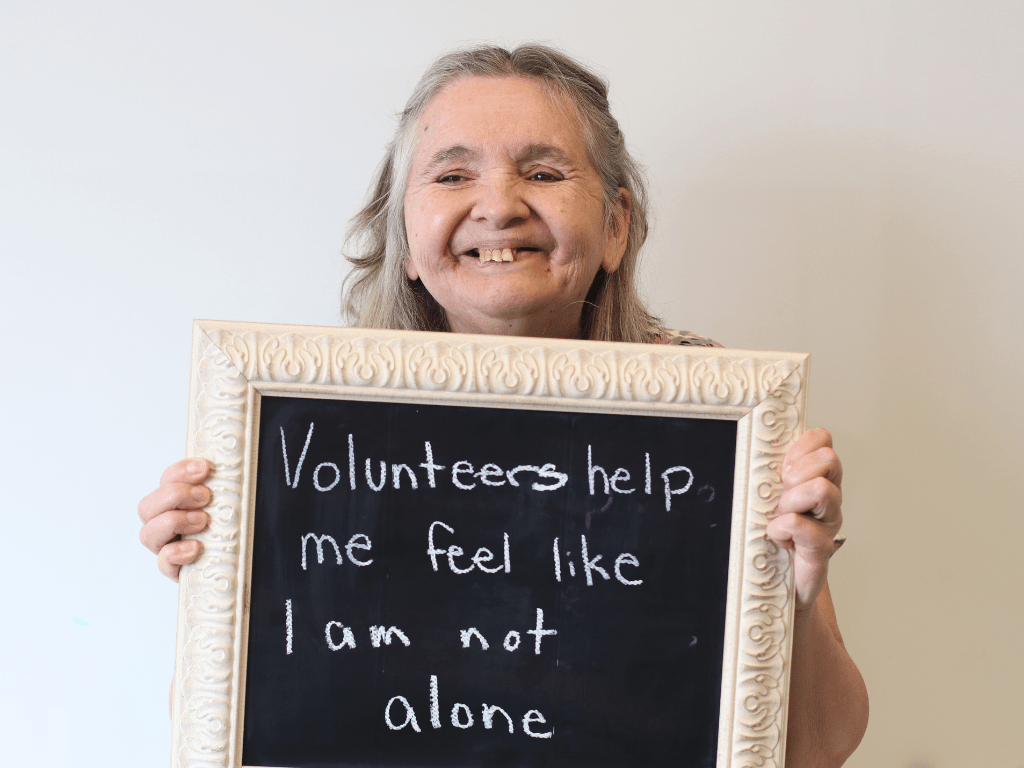 A lady holding a chalk board that says "Volunteers help me feel like I am not alone"