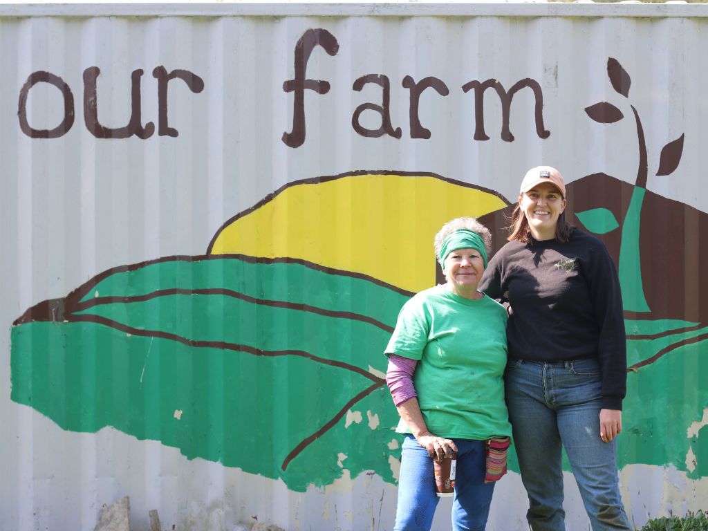 Our Farm Coordinator Jenny with Our Farm Agricultural Farmer Laura smiling beside the Our Farm mural outside the farm shed.