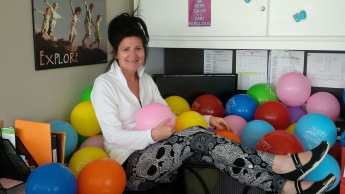 Carol from Early Learning smiling with a bunch of colourful Happy Birthday balloons at her desk.
