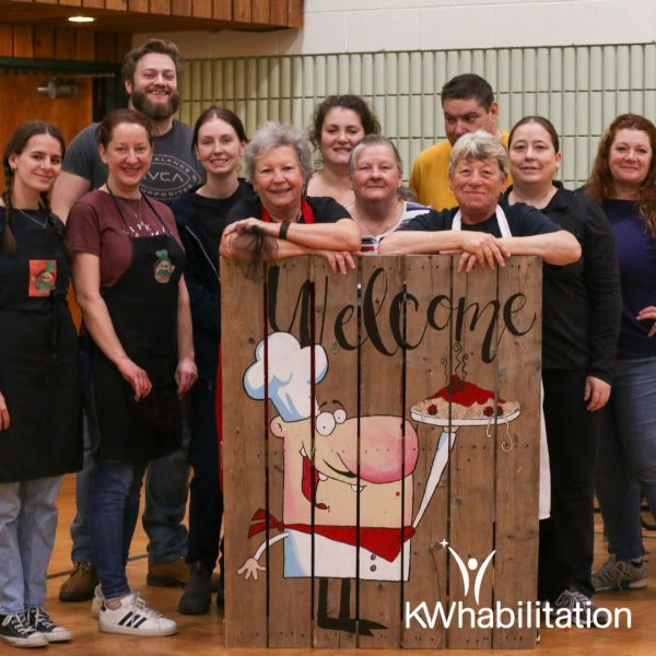 Committee For Excellence smiling beside our hand painted wooden welcome sign at the Annual 2024 Spaghetti Dinner. On the sign is a chef holding a plate of spaghetti.