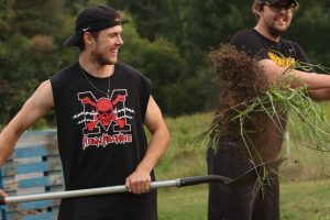 Two students from the University of Waterloo volunteering on Our Farm. Student on left is tossing soil in the air with a shovel while smiling. Student behind on the right is watching them and smiling.